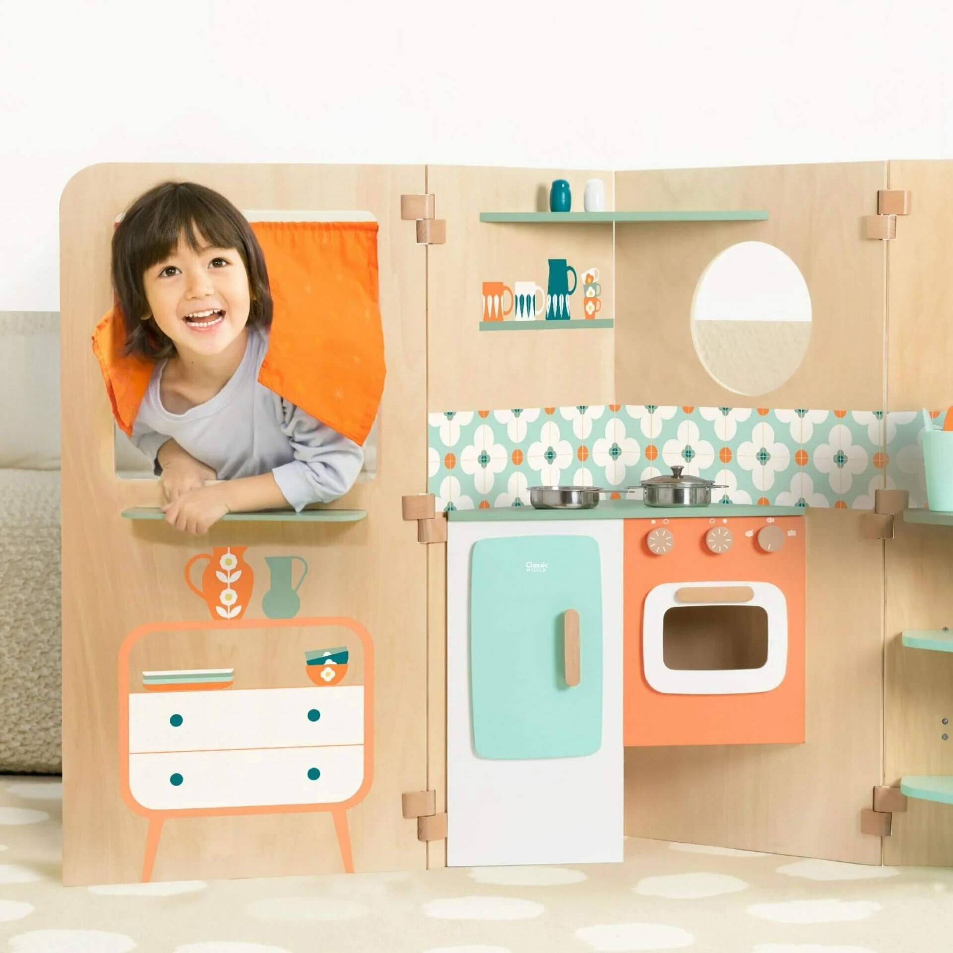 A child smiling through the orange curtained window of the My Little House, with a colourful kitchen featuring an oven, fridge, and shelves.