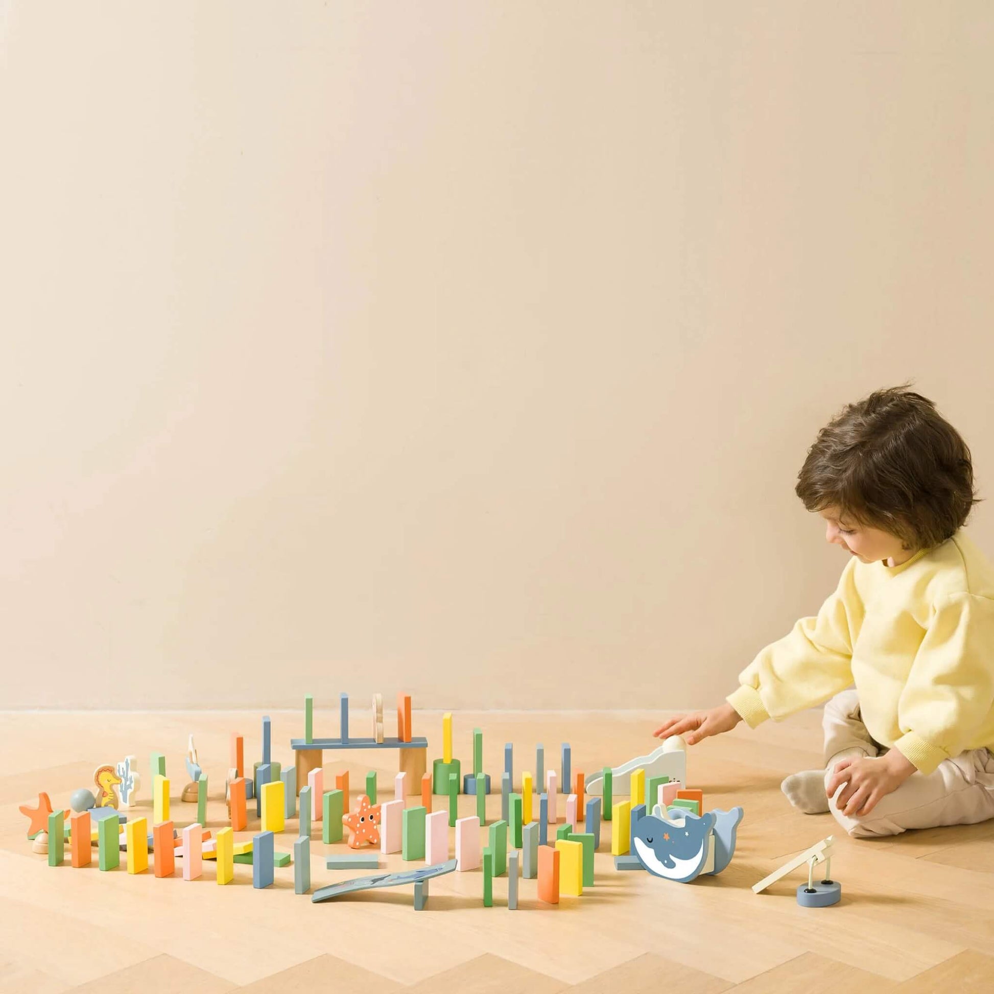 Child sitting on a wooden floor, fully engaged in setting up the colourful ocean domino pieces and accessories in a large pattern.