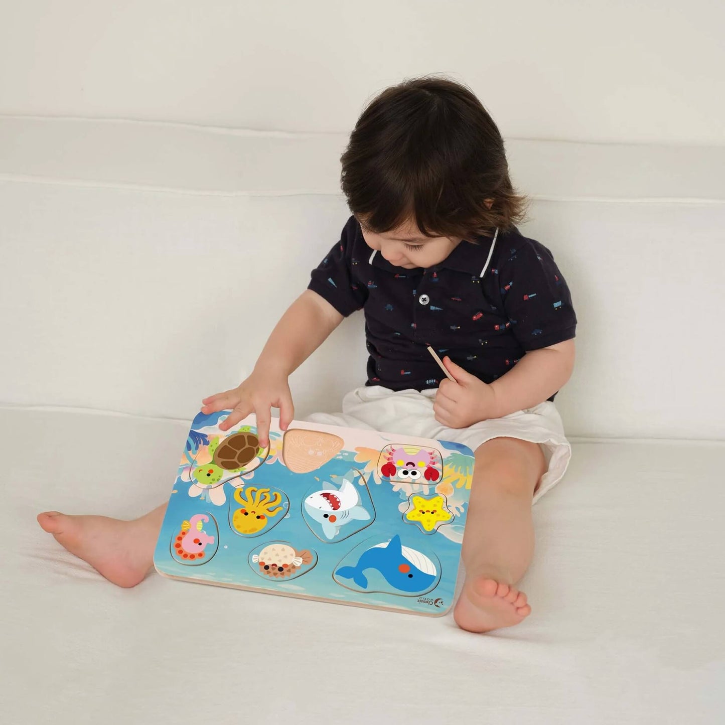 Toddler sitting on a light-coloured sofa, playing with a colourful wooden ocean puzzle, placing a turtle piece into the matching outline on the puzzle board.