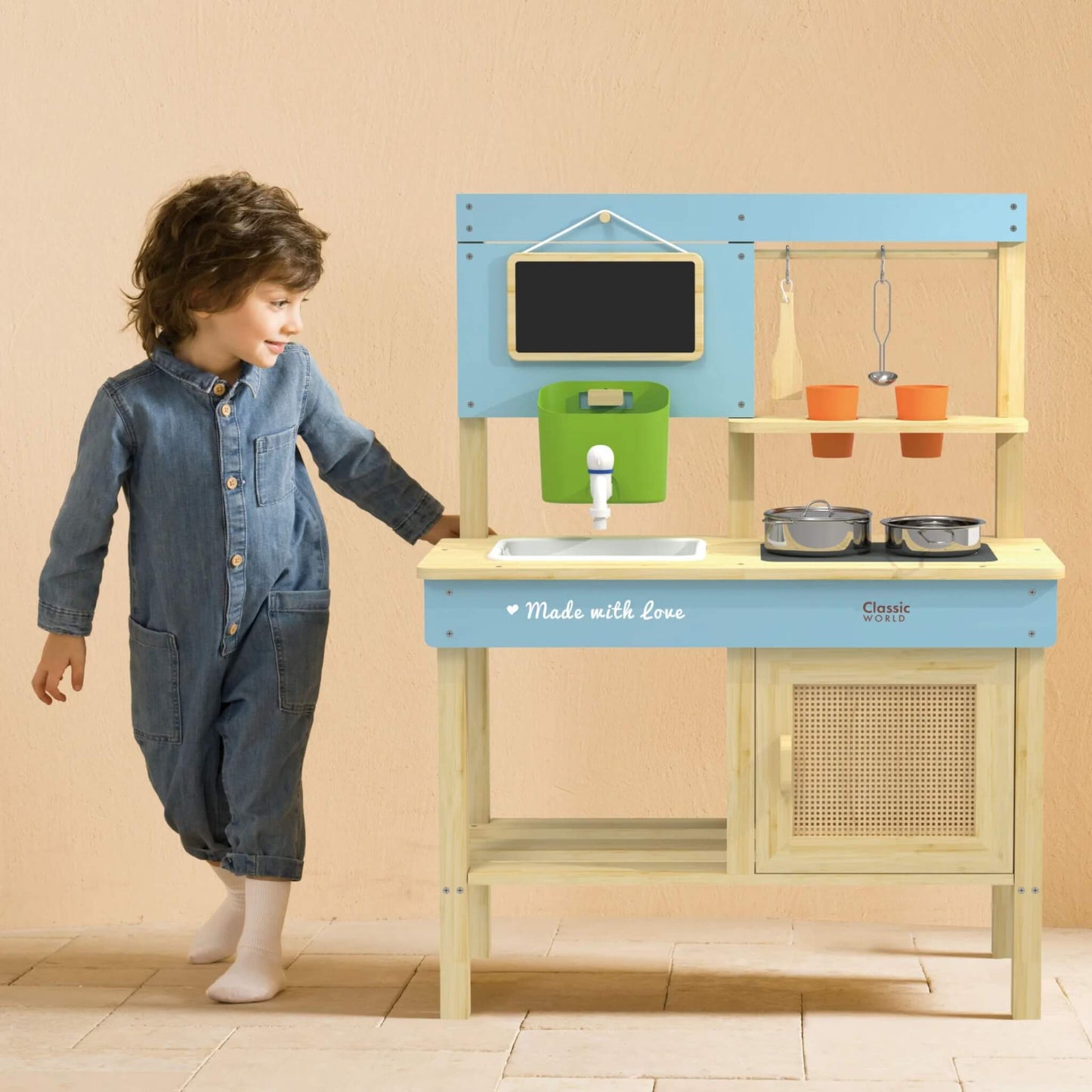 Young child wearing a denim jumpsuit standing next to a toy wooden outdoor kitchen with blue panels, a blackboard, working sink, hanging utensils, and metal pots.