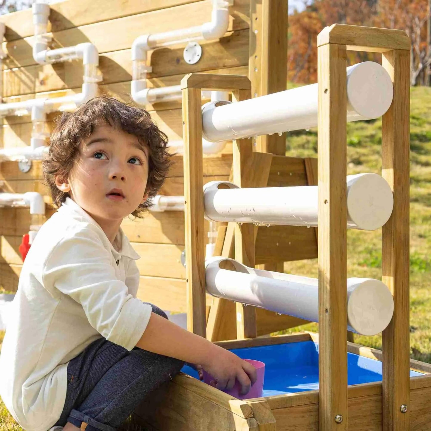 Young child playing with a wooden outdoor water play wall, catching water in a pink cup as it flows through white chutes into a blue basin.