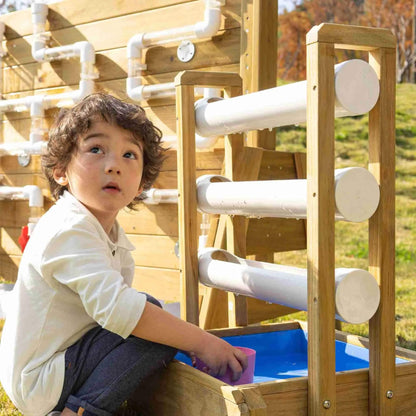 Young child playing with a wooden outdoor water play wall, catching water in a pink cup as it flows through white chutes into a blue basin.