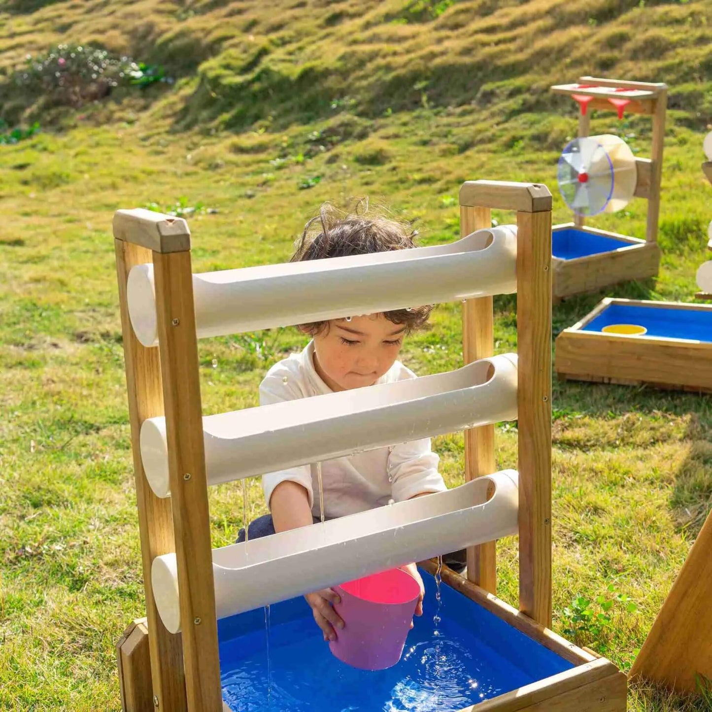 Child scooping water from an outdoor water play setup, watching streams pour down chutes into a blue basin on a grassy play area.