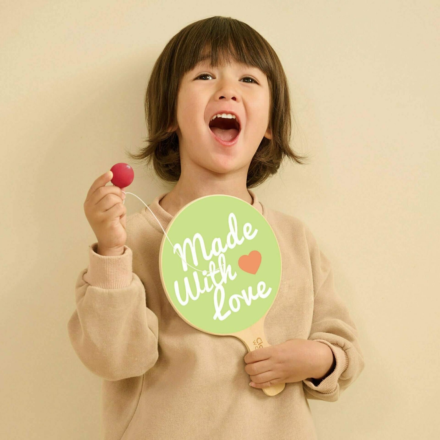 A young child smiling and holding a wooden paddle ball toy in one hand and the attached red ball in the other, with the green paddle face showing playful printed text.