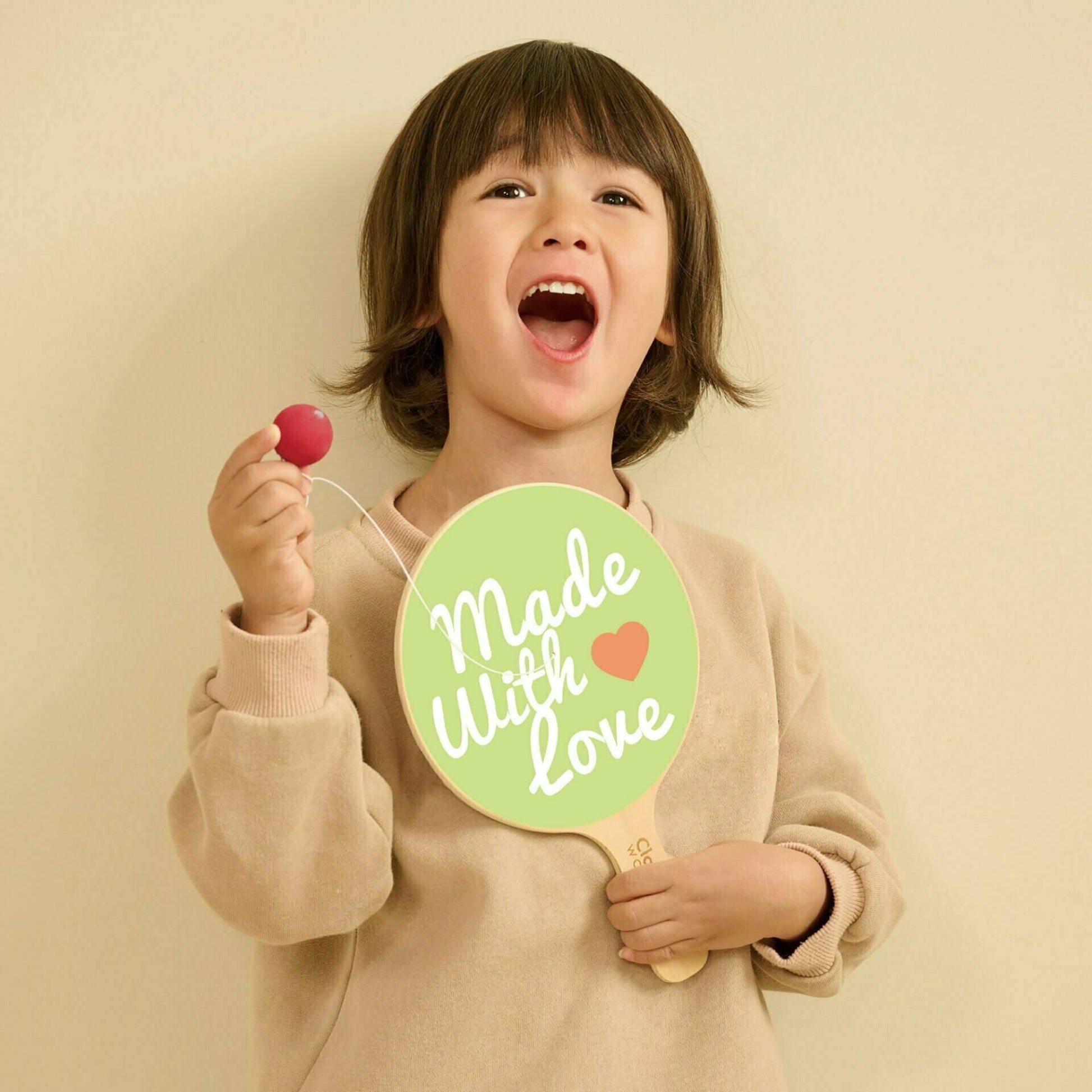 A young child smiling and holding a wooden paddle ball toy in one hand and the attached red ball in the other, with the green paddle face showing playful printed text.