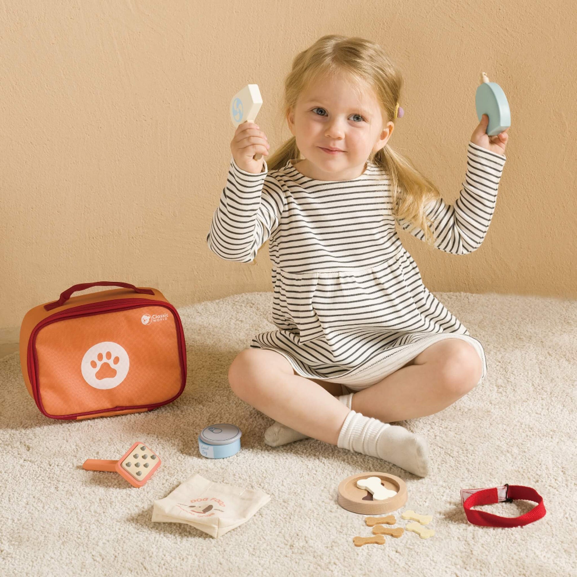 Child sitting on carpet playing with wooden pet care toys, holding grooming tools beside orange storage case.