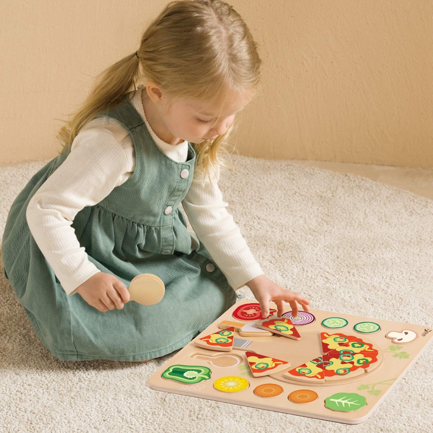 Young child playing with a wooden pizza puzzle toy, placing toppings and holding a wooden utensil on a soft rug.