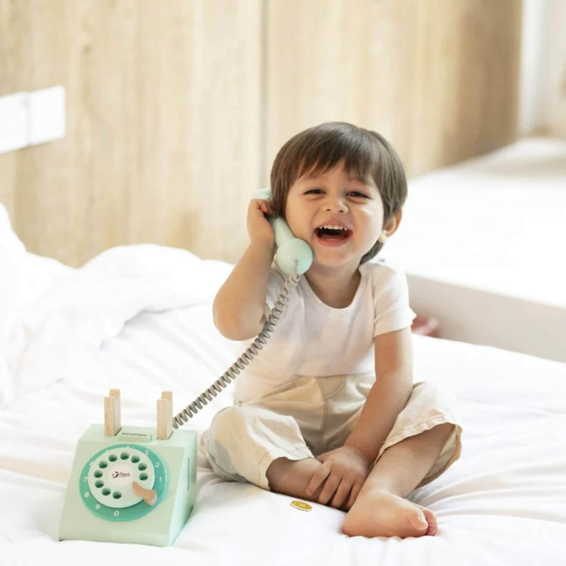 A young child sitting on a bed, smiling while holding a pale turquoise toy telephone handset to their ear, with the toy base resting beside them.