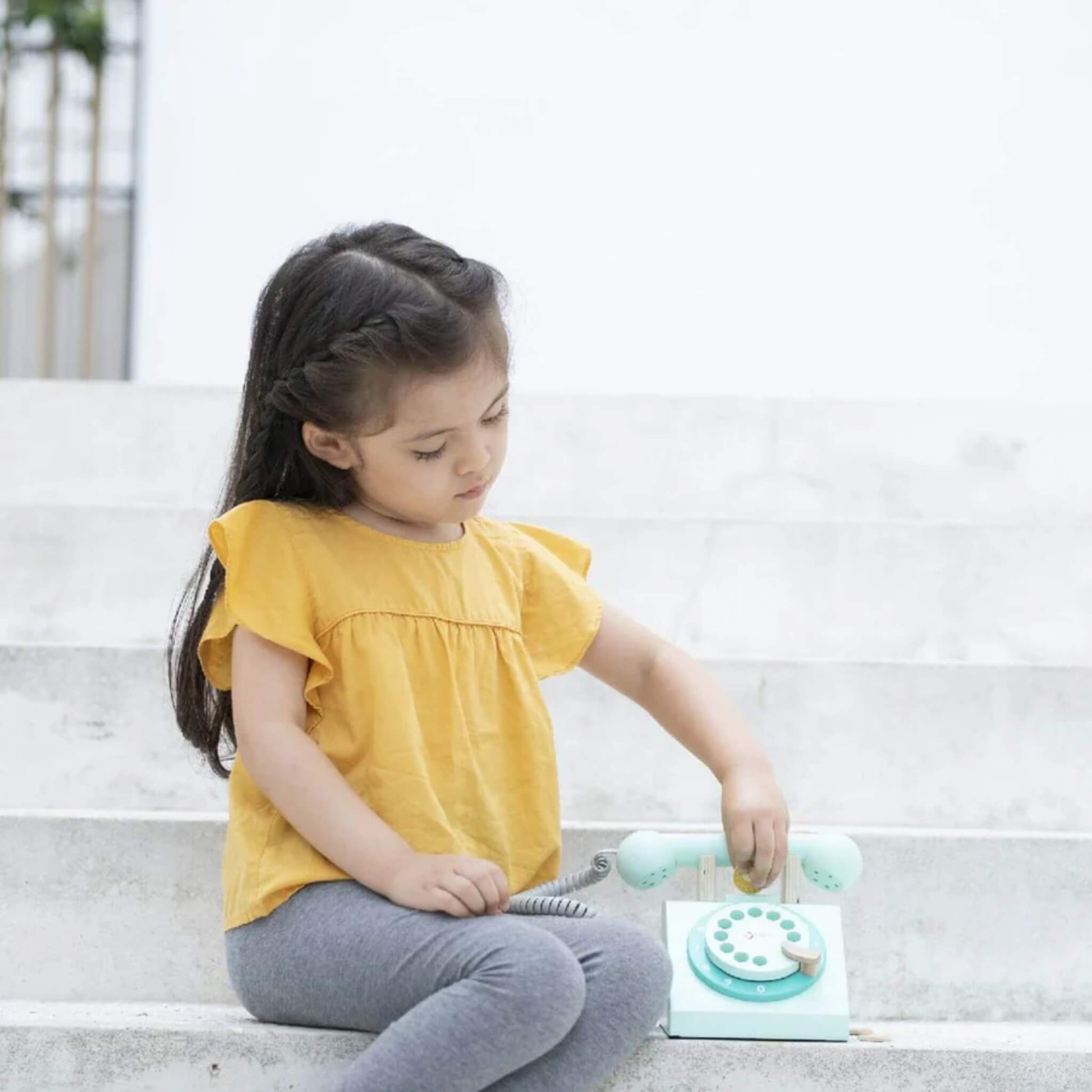 A child in a mustard yellow top sitting on outdoor steps, placing a coin into the slot of a wooden toy telephone with a rotary dial.