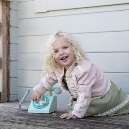 A blonde-haired child kneeling on a wooden deck, smiling while turning the rotary dial on a pale turquoise toy telephone.