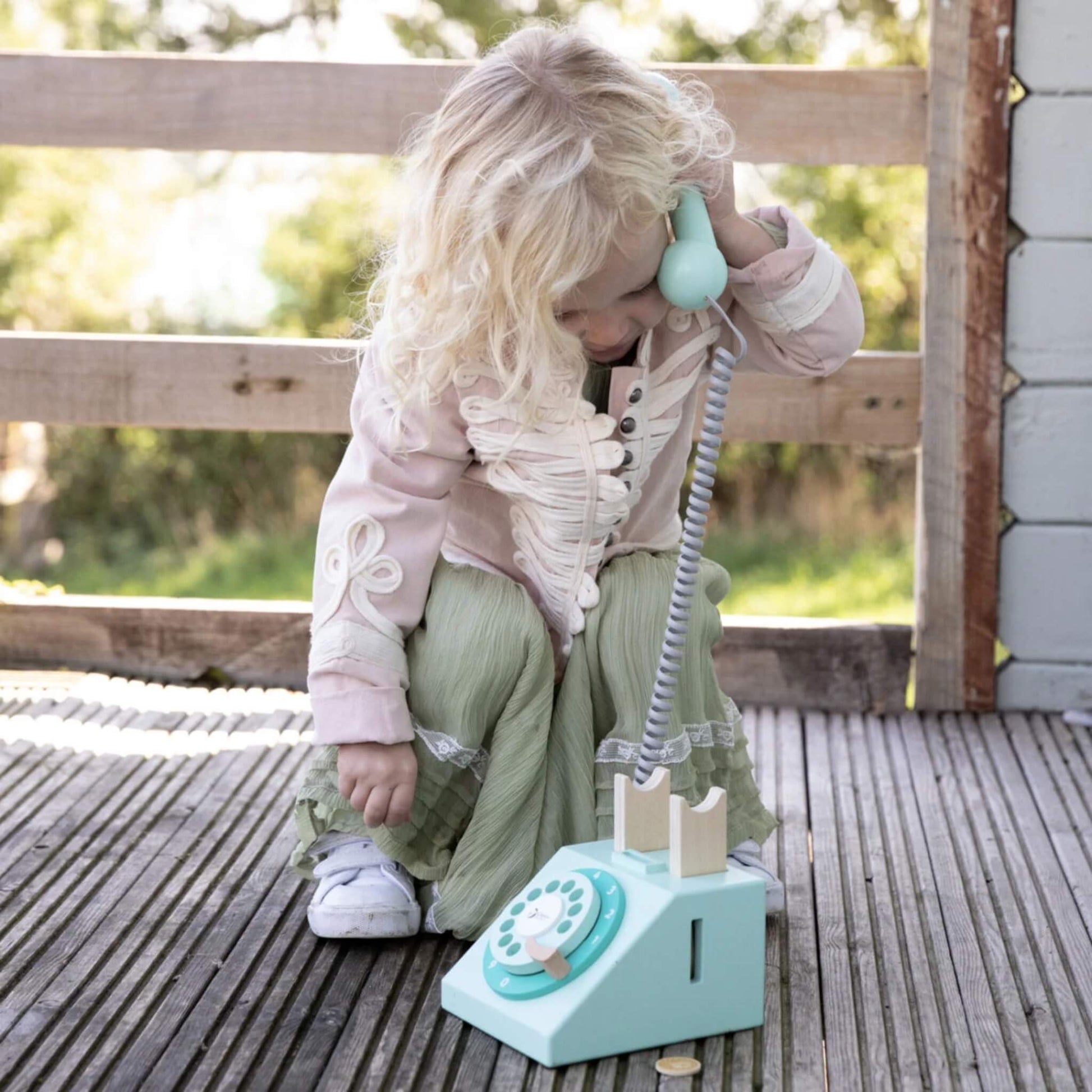 A child crouching on a wooden deck, holding the handset of a toy telephone to their ear while the phone base rests nearby.