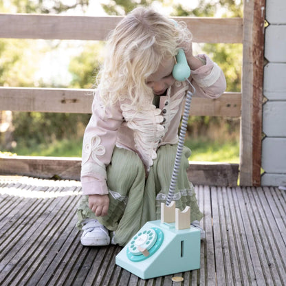 A child crouching on a wooden deck, holding the handset of a toy telephone to their ear while the phone base rests nearby.