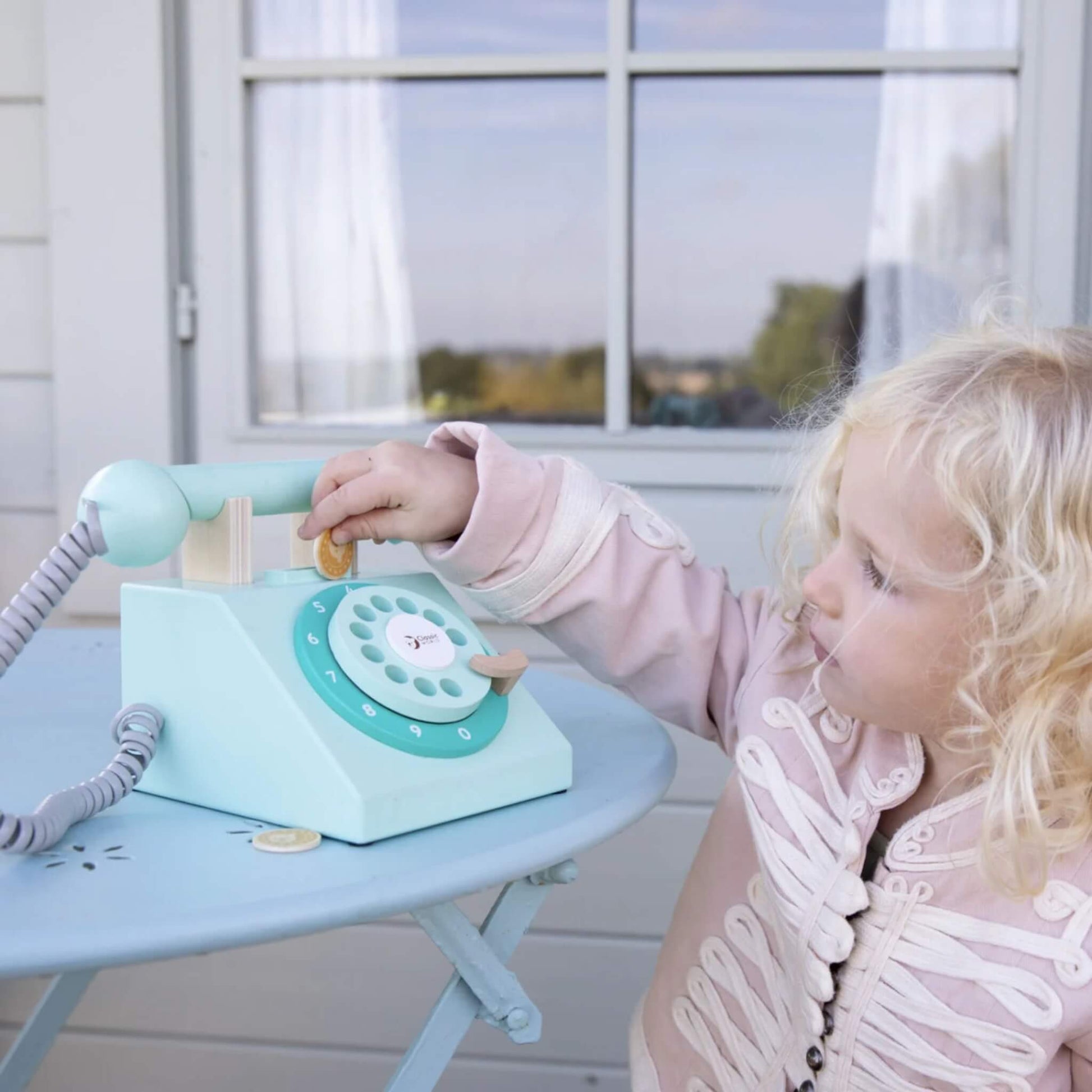 A child standing next to a round blue table, dropping a gold play coin into the slot of a wooden toy telephone with a rotary dial.