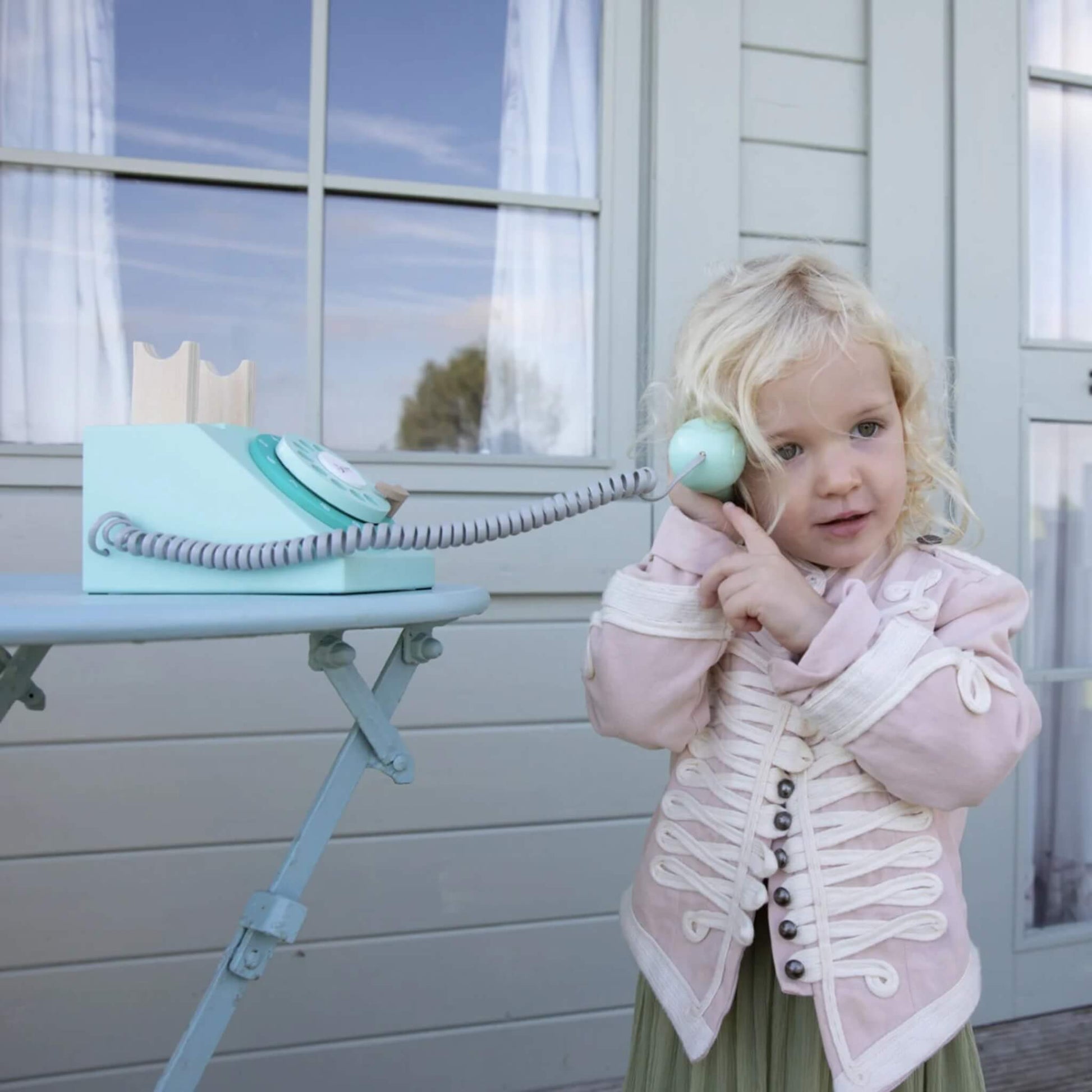 A child holding a toy phone handset to their ear with one hand and gesturing with the other, standing beside a small round table with the phone base on top.