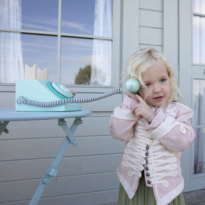 A child holding a toy phone handset to their ear with one hand and gesturing with the other, standing beside a small round table with the phone base on top.
