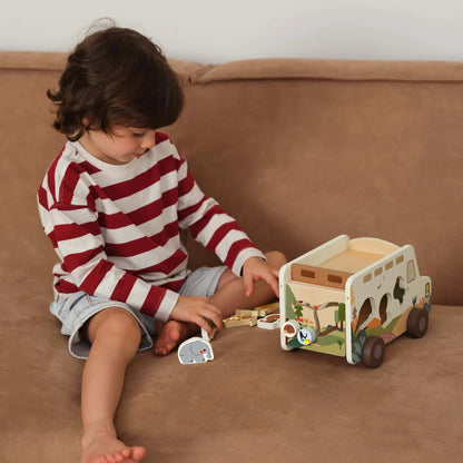 A toddler sitting on a tan sofa, playing with the safari truck and wooden animal figures, placing them into the matching shaped cut-outs.