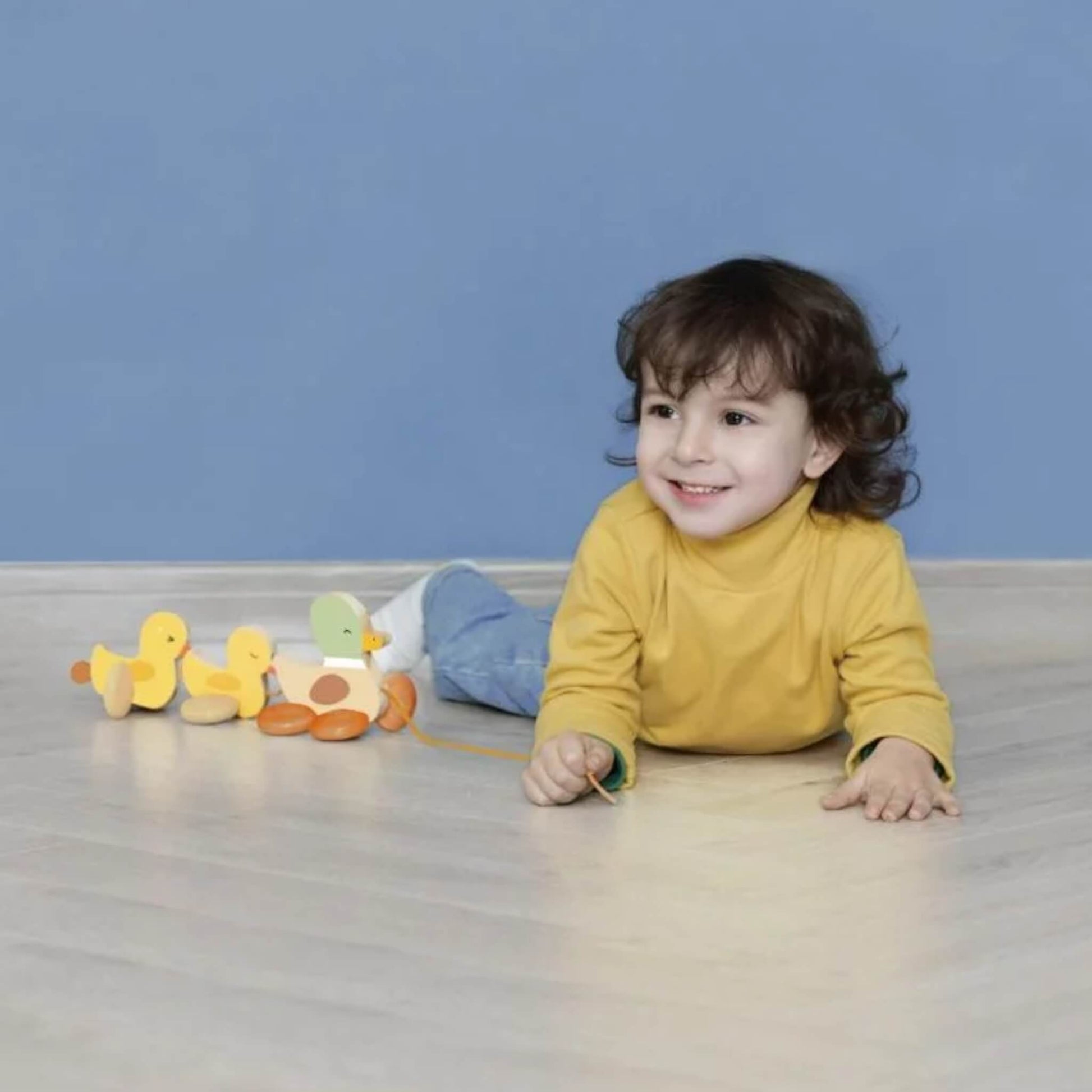 A smiling child lying on a wooden floor, holding the string attached to a pull-along duck family toy placed beside them.