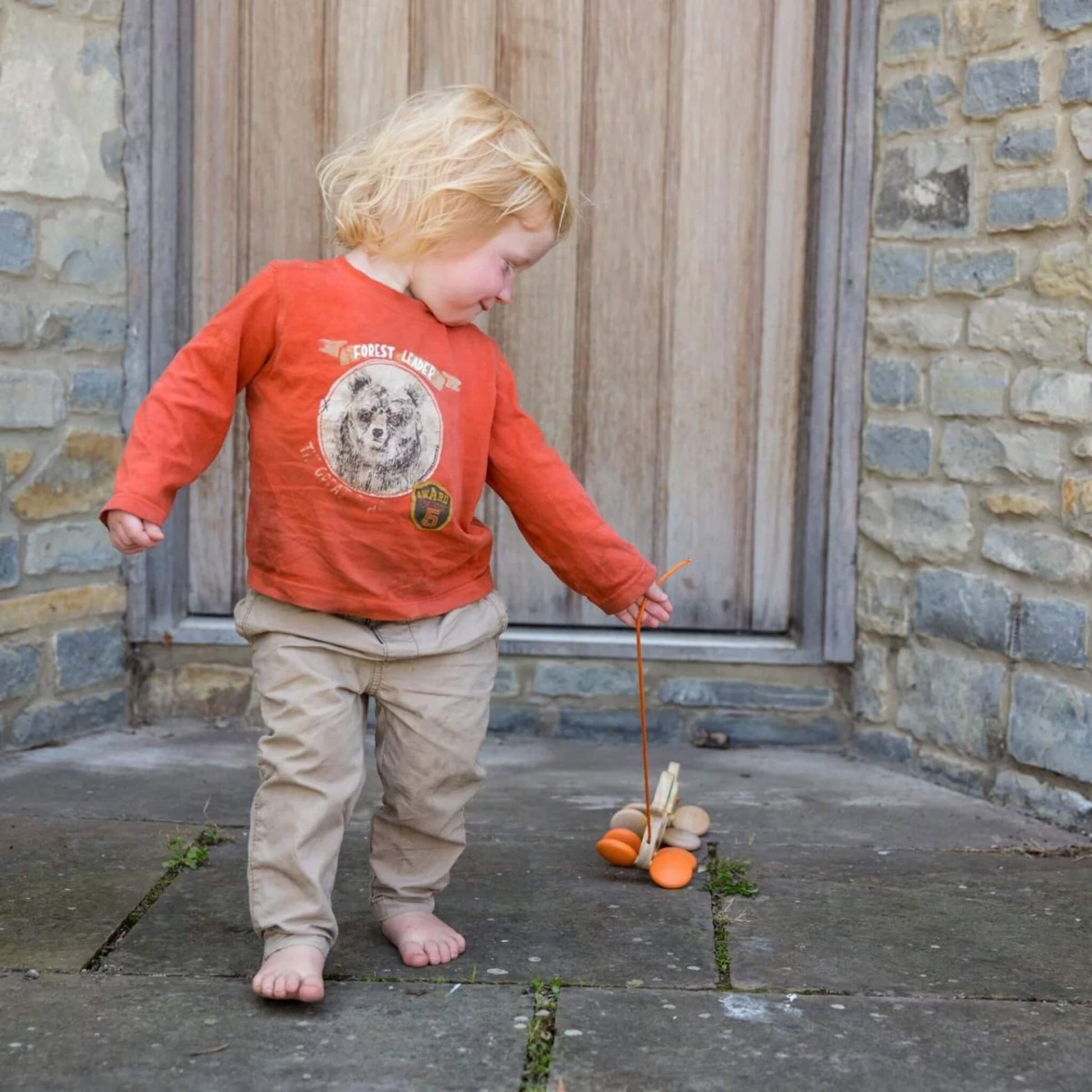 A barefoot toddler outdoors in front of a stone building, pulling a wooden duck family toy by its orange string.