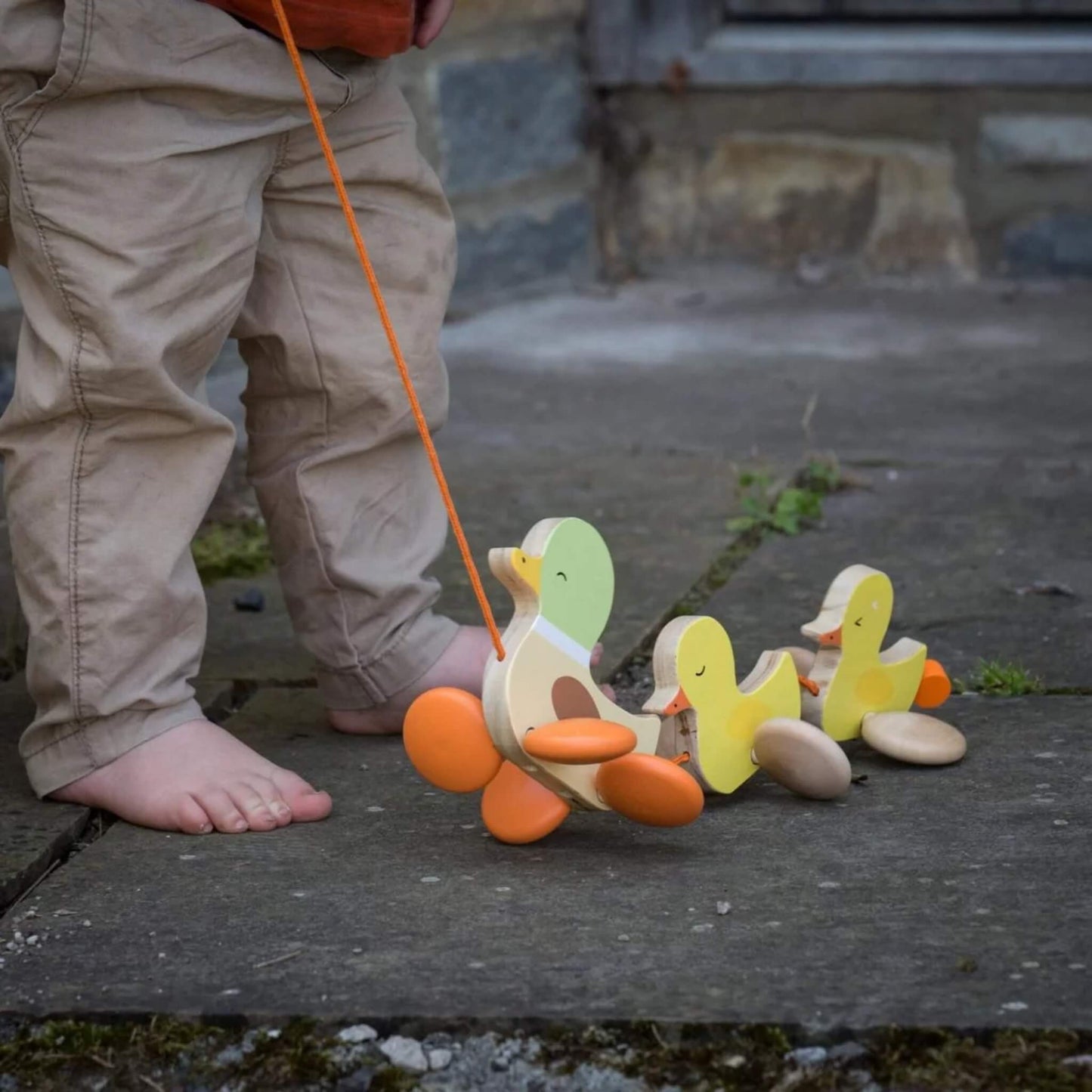 A close-up of the wooden pull-along duck family toy on a stone path, being held by a child in beige trousers.