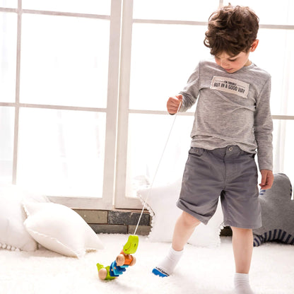 Young child walking barefoot on a fluffy white carpet, pulling a colourful wooden toy crocodile by a string in a sunlit room.