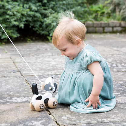 A toddler in a soft green dress sitting on a stone patio, smiling while looking at a wooden pull-along dog toy with spotted wheels and floppy ears.