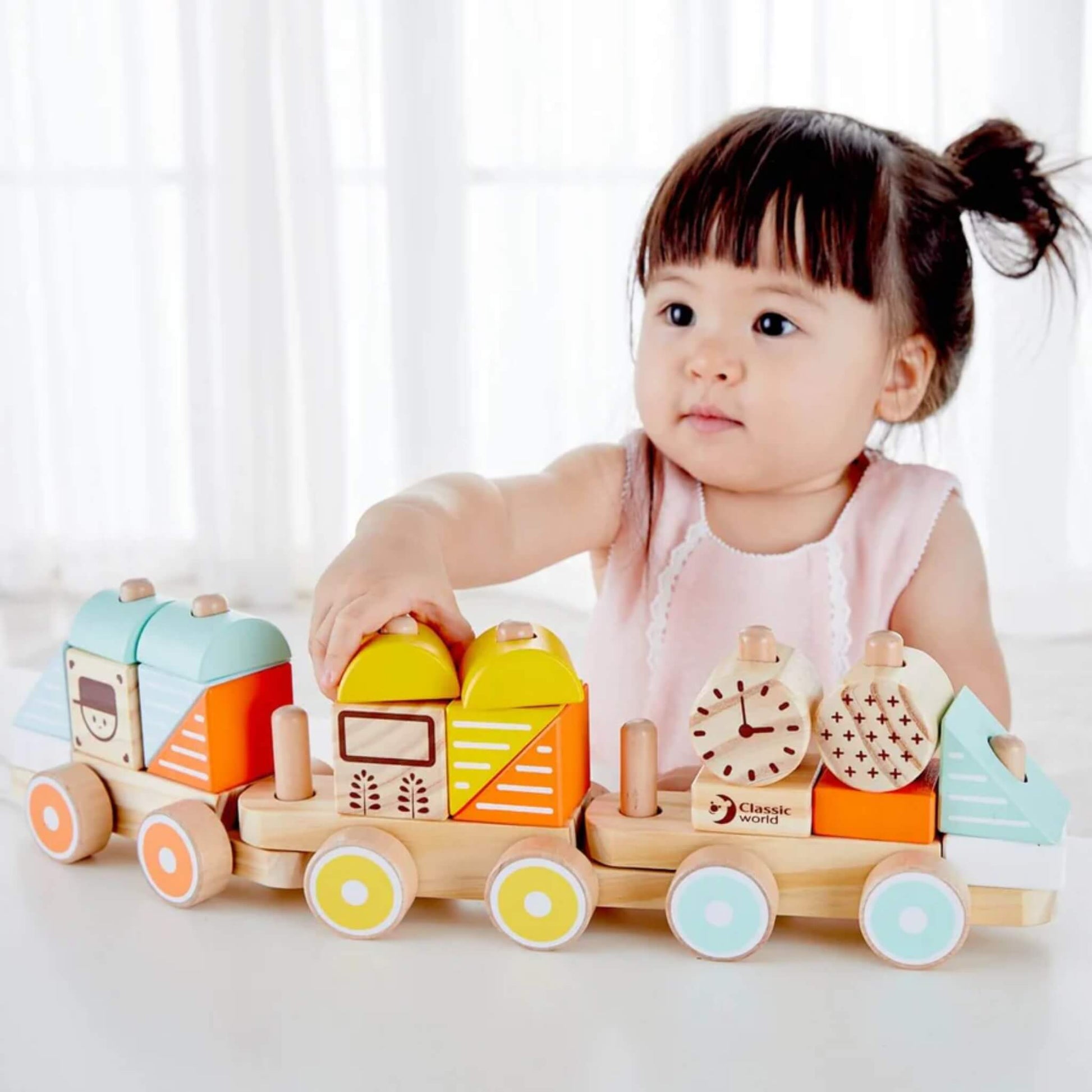 A toddler in a pale pink dress arranges wooden blocks onto the pull-along train, focusing intently at a table near a bright window.