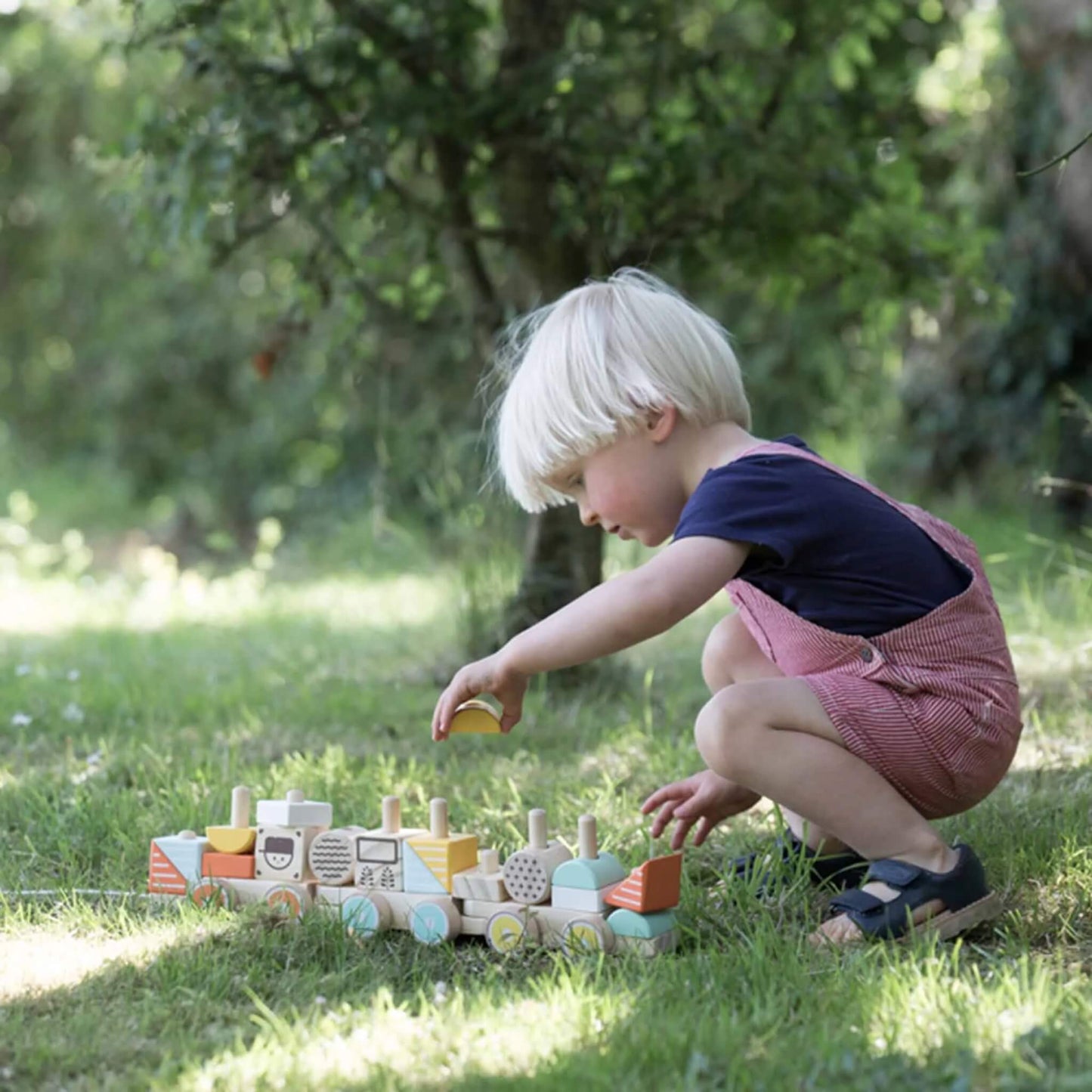 A young child crouches outside on the grass, carefully placing a yellow block onto a wooden stacking train’s peg.