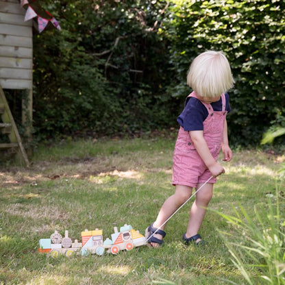 A toddler in red dungarees pulls an assembled wooden pull-along train by its white string along a sunlit patch of garden.