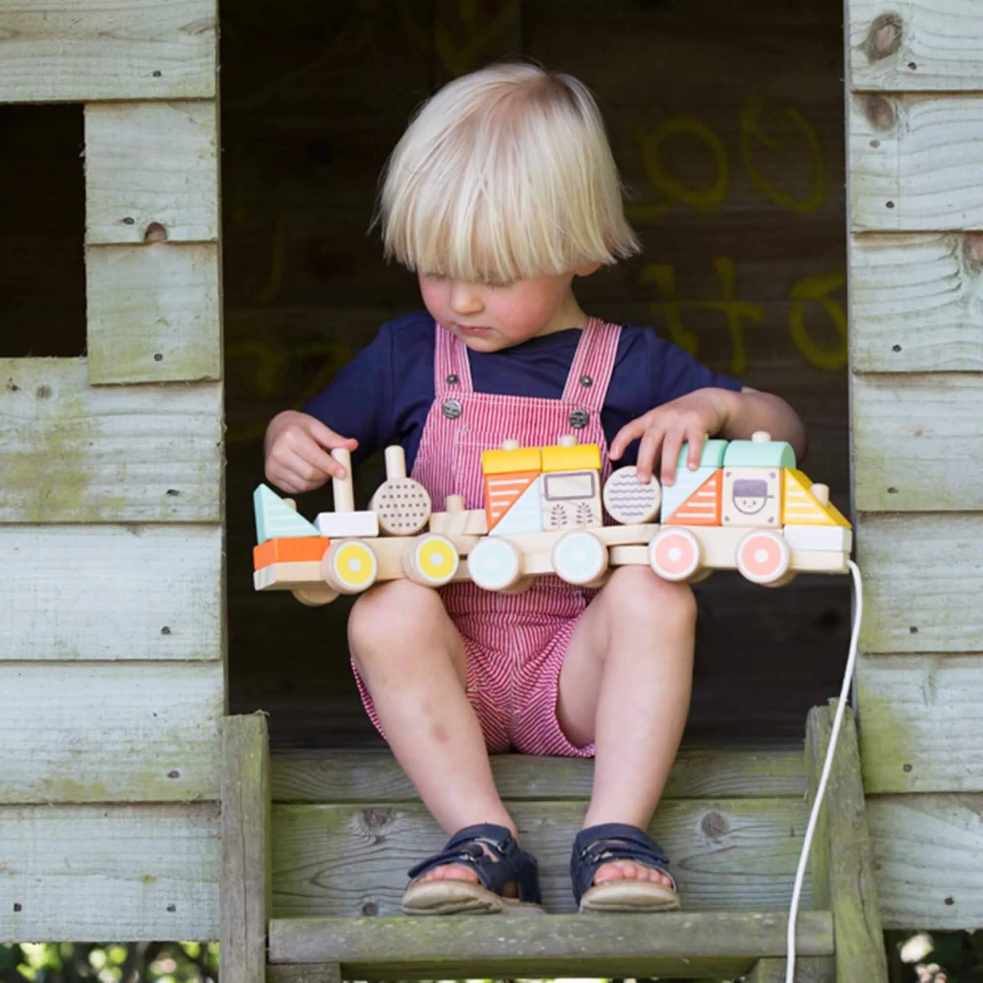 A small child sits in a wooden playhouse, balancing an assembled stacking train on their lap while exploring its different shapes.