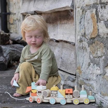 A child sitting beside a rustic stone wall plays quietly with individual train blocks, rotating them in their hands and placing them on the train.
