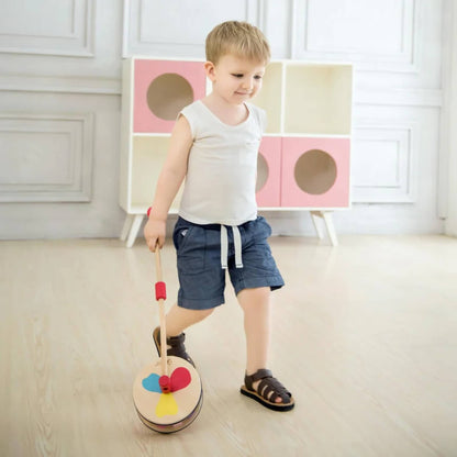 Toddler in blue shorts and sandals smiling while walking indoors, pushing a colourful wooden push toy with a propeller design.