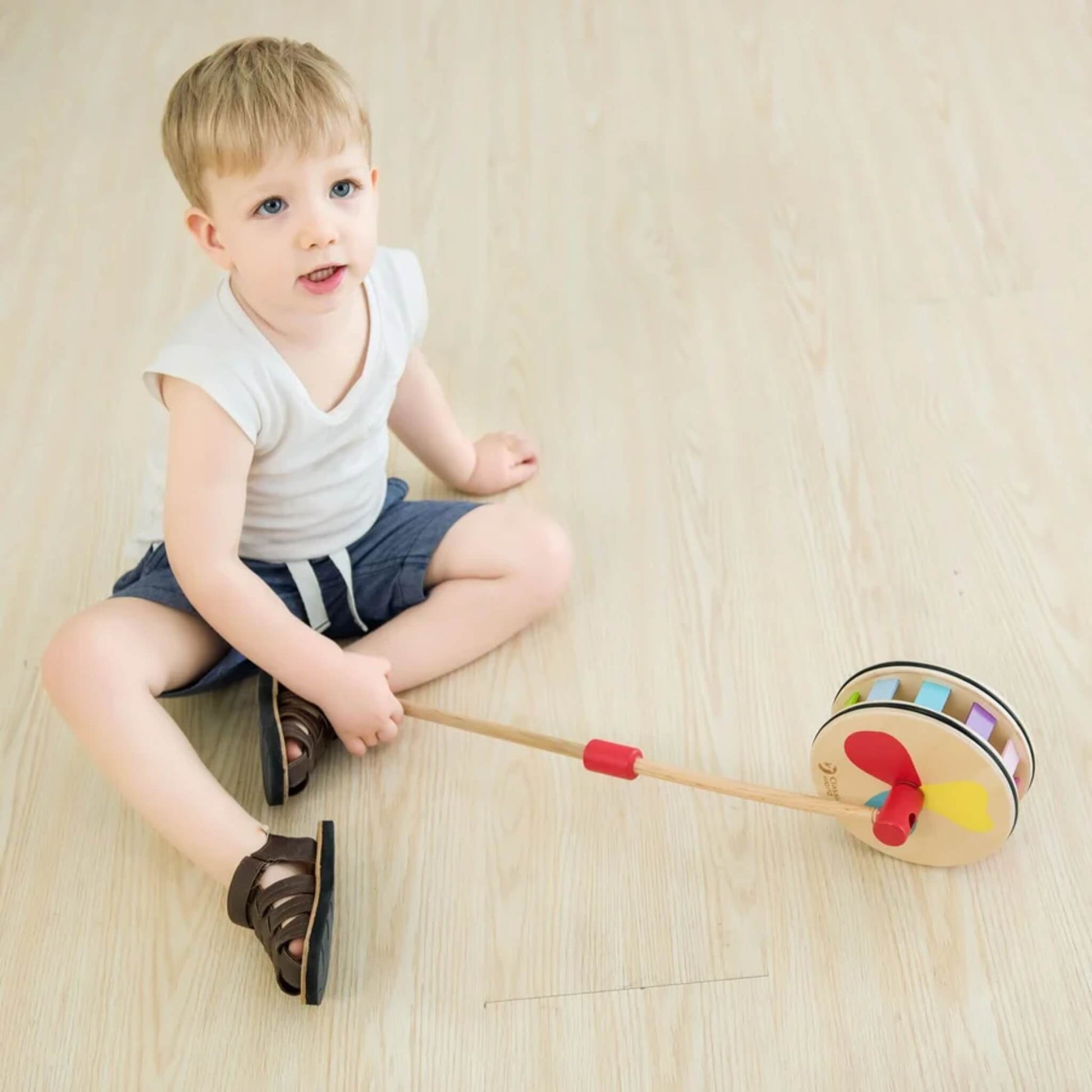 Young child sitting on a wooden floor, holding a rainbow push toy with bright paddles and curved wooden sides.
