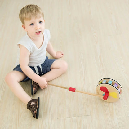 Young child sitting on a wooden floor, holding a rainbow push toy with bright paddles and curved wooden sides.