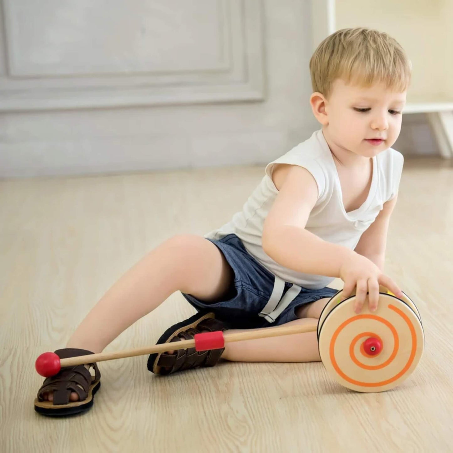 Child in a white top seated and playing with a wooden rolling toy featuring an orange spiral on the side.