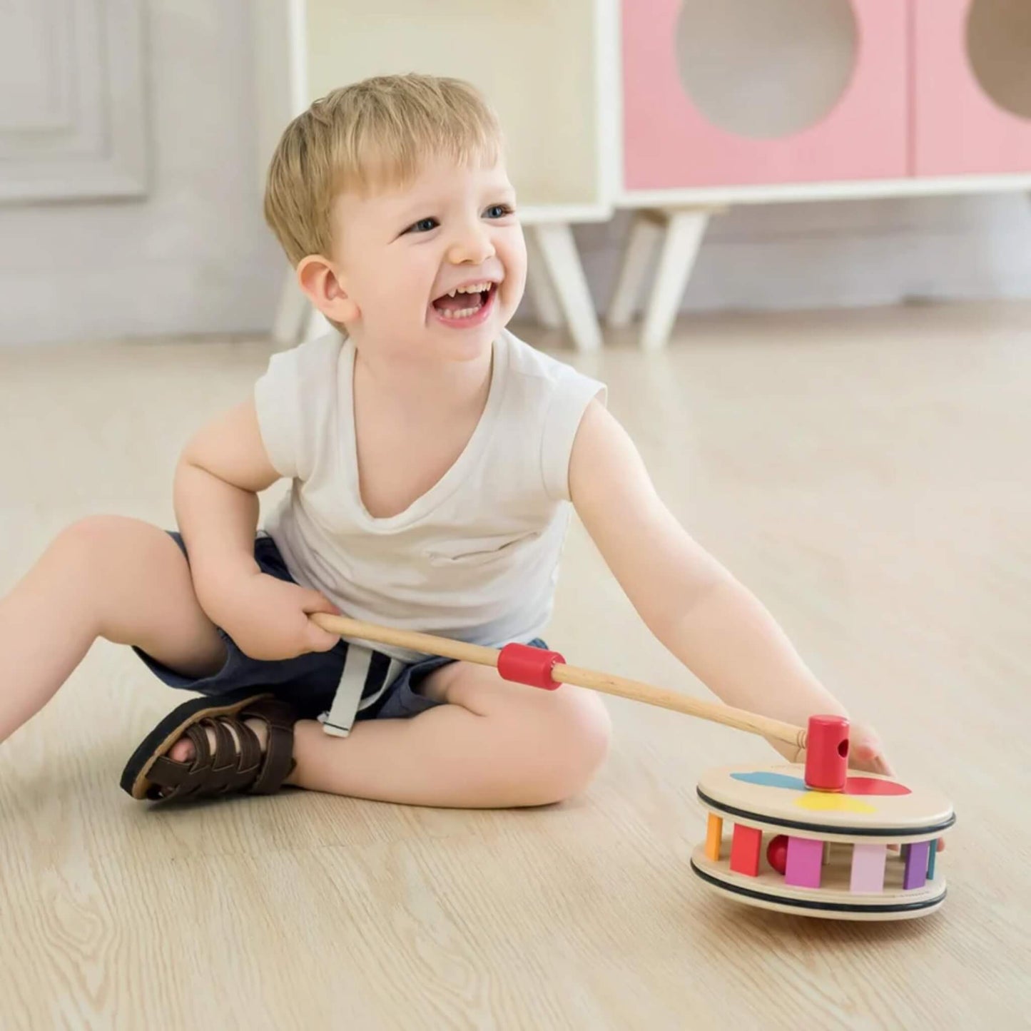 Toddler grinning and holding a rainbow push toy while sitting on the floor, surrounded by bright natural light.