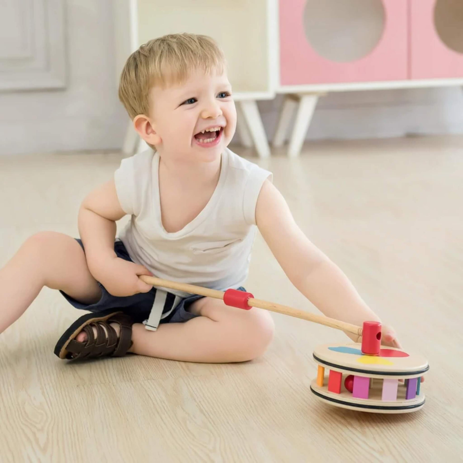 Toddler grinning and holding a rainbow push toy while sitting on the floor, surrounded by bright natural light.