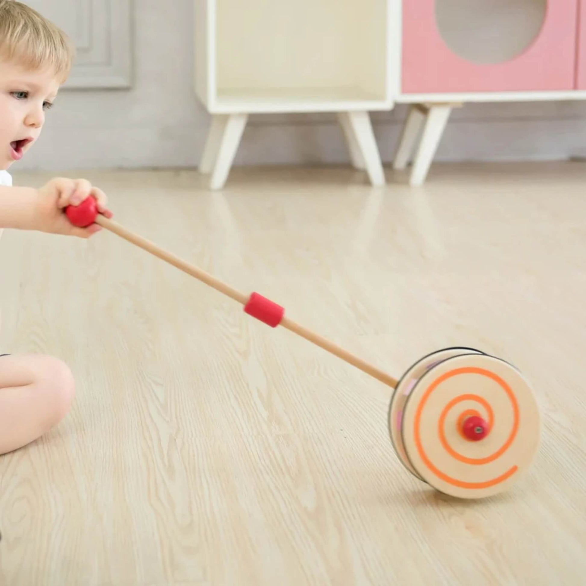 Child leaning forward, pushing a wooden push toy with an orange swirl on the side and visible inner paddles.
