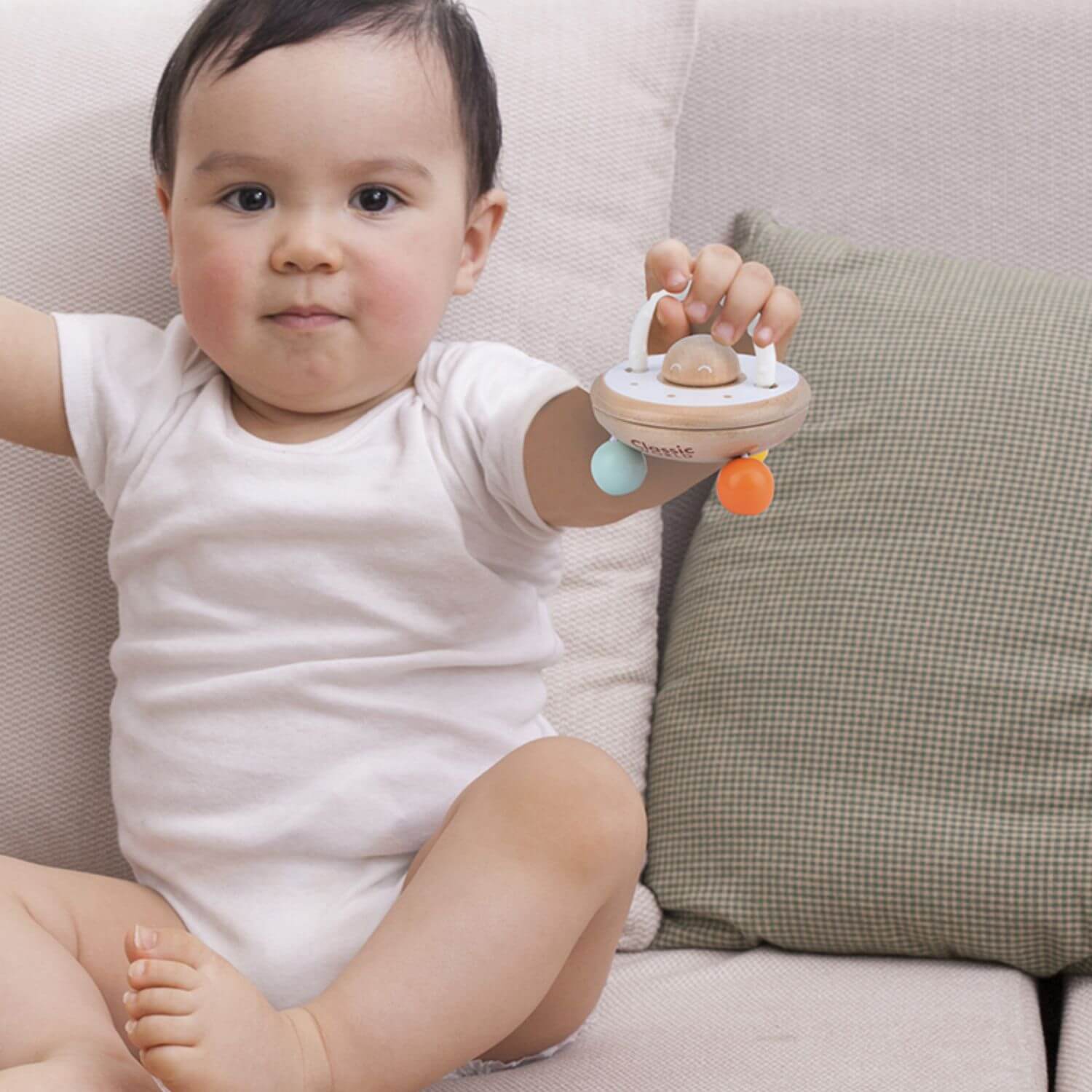 A baby sitting on a sofa holding a U.F.O shaped wooden rattle, showcasing its easy-to-grasp design and engaging features for little hands.
