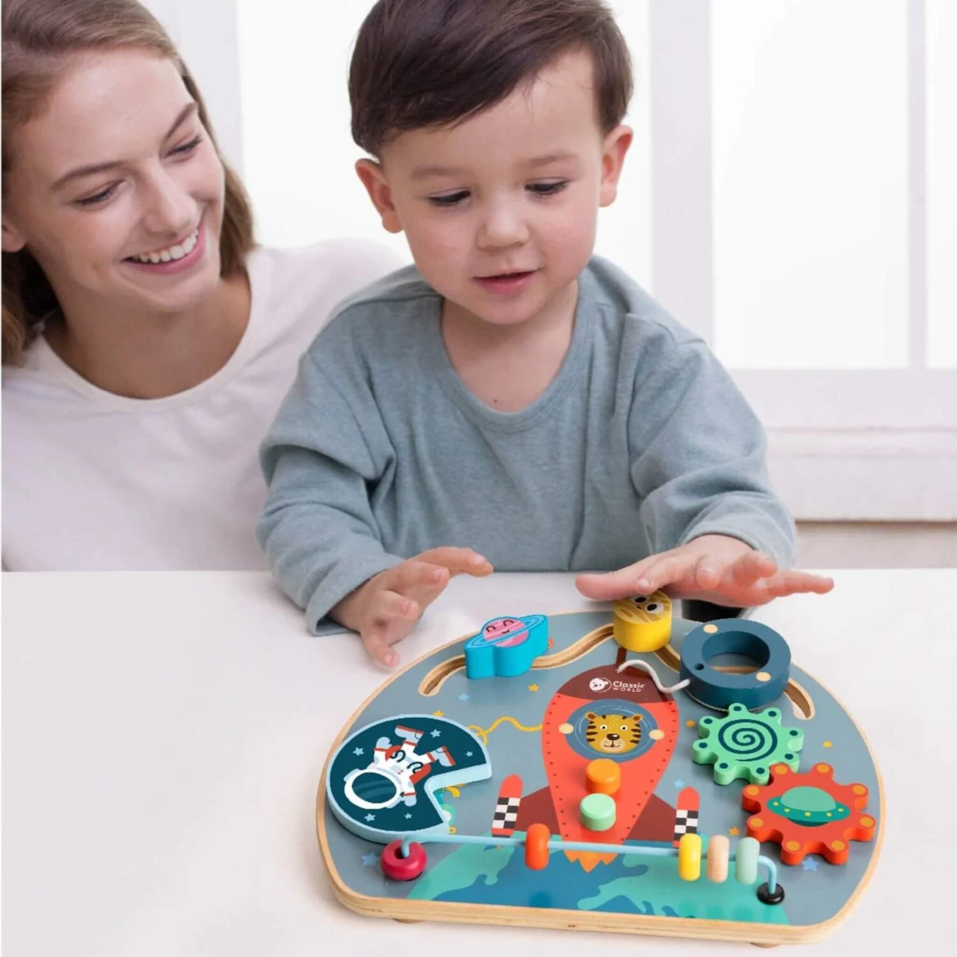 Child smiling and playing at a table with a space-themed wooden busy board, interacting with the elements.