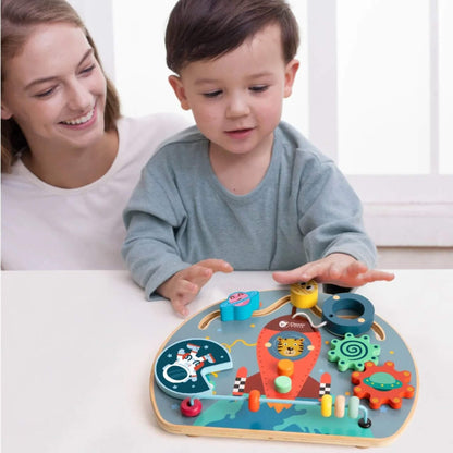 Child smiling and playing at a table with a space-themed wooden busy board, interacting with the elements.