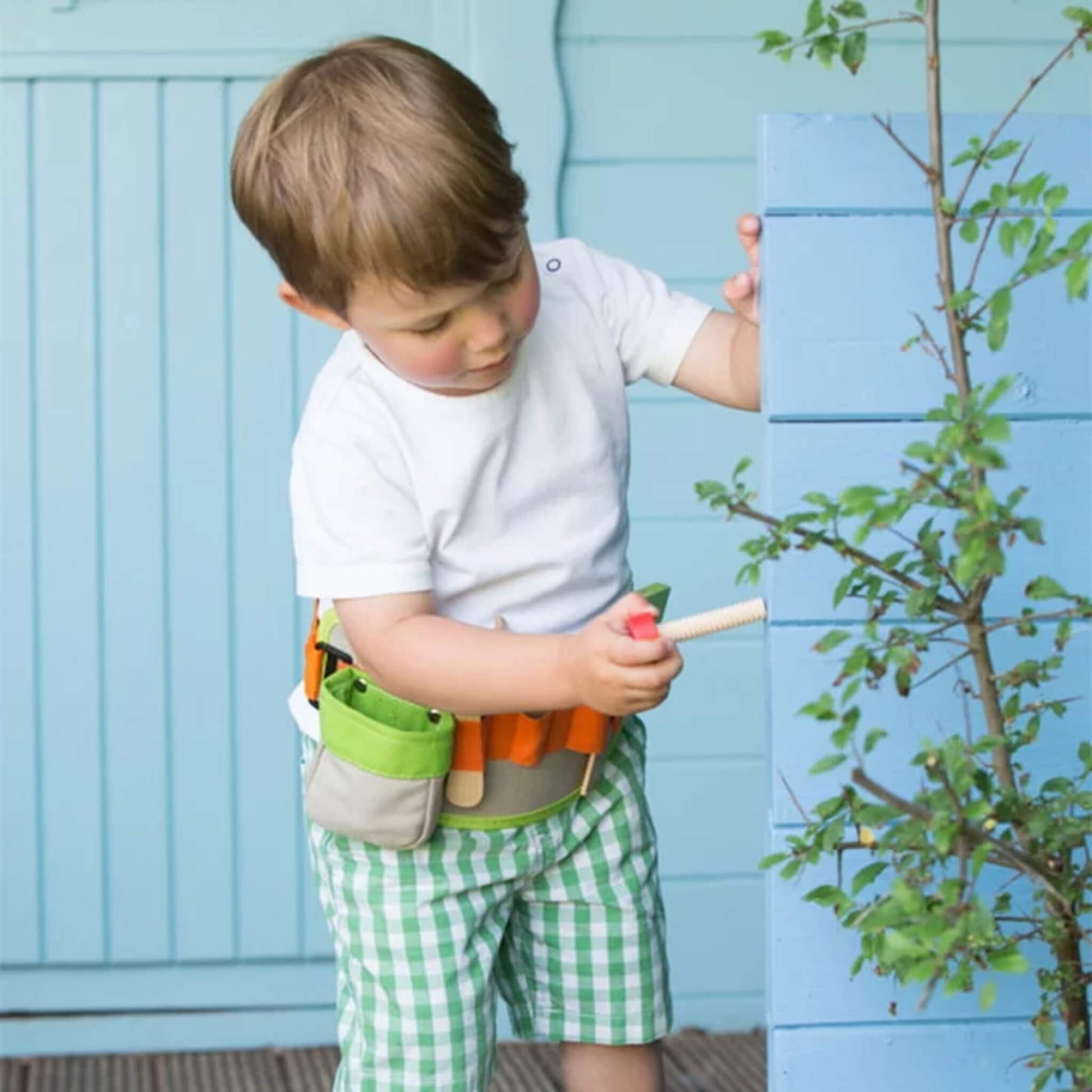 A young child wearing a tool belt uses a wooden screwdriver to pretend-fix a light blue fence, with a small leafy plant growing nearby.