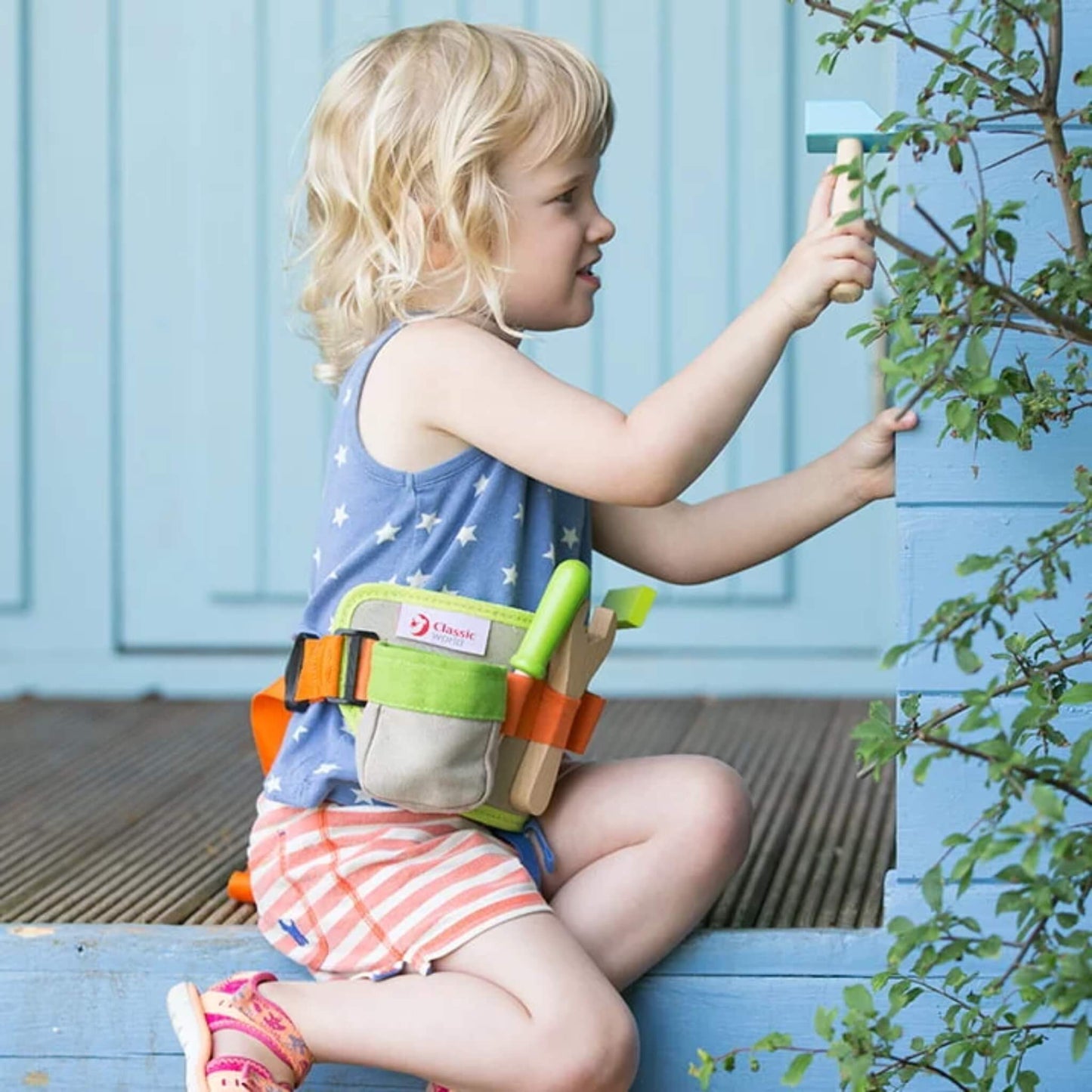 A little girl in a star-print top and striped shorts kneels next to a blue wooden step, using a wooden hammer while wearing a tool belt with green and orange trim.