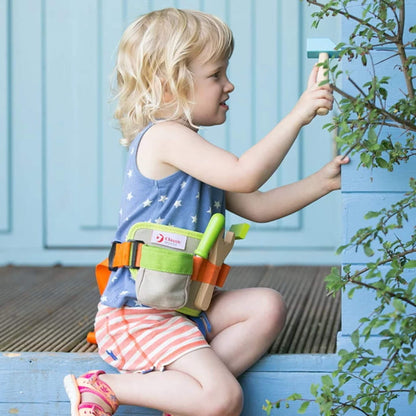 A little girl in a star-print top and striped shorts kneels next to a blue wooden step, using a wooden hammer while wearing a tool belt with green and orange trim.