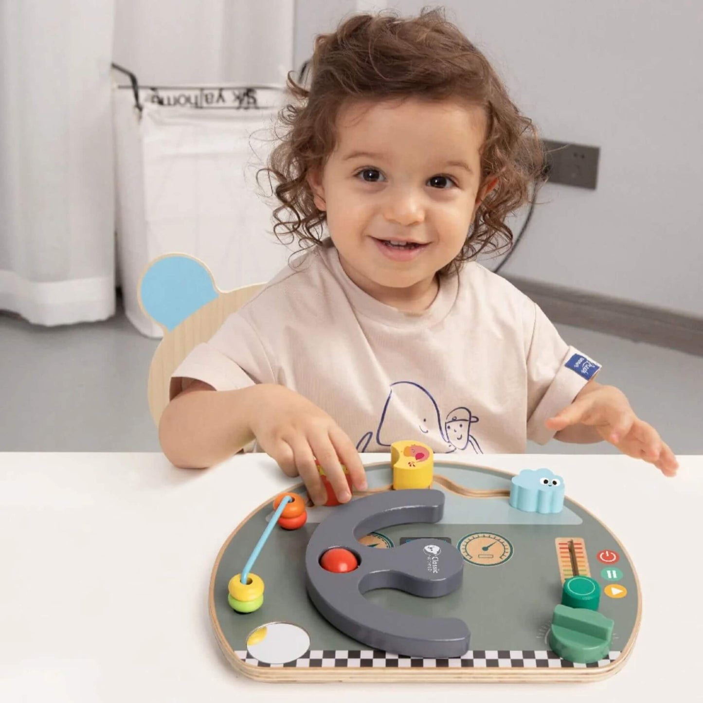 Smiling toddler playing at a table with a wooden busy board, hands on the activity pieces.
