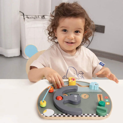 Smiling toddler playing at a table with a wooden busy board, hands on the activity pieces.