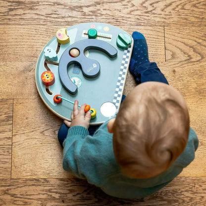 Overhead view of a young child sitting on the floor, exploring a busy board with both hands.