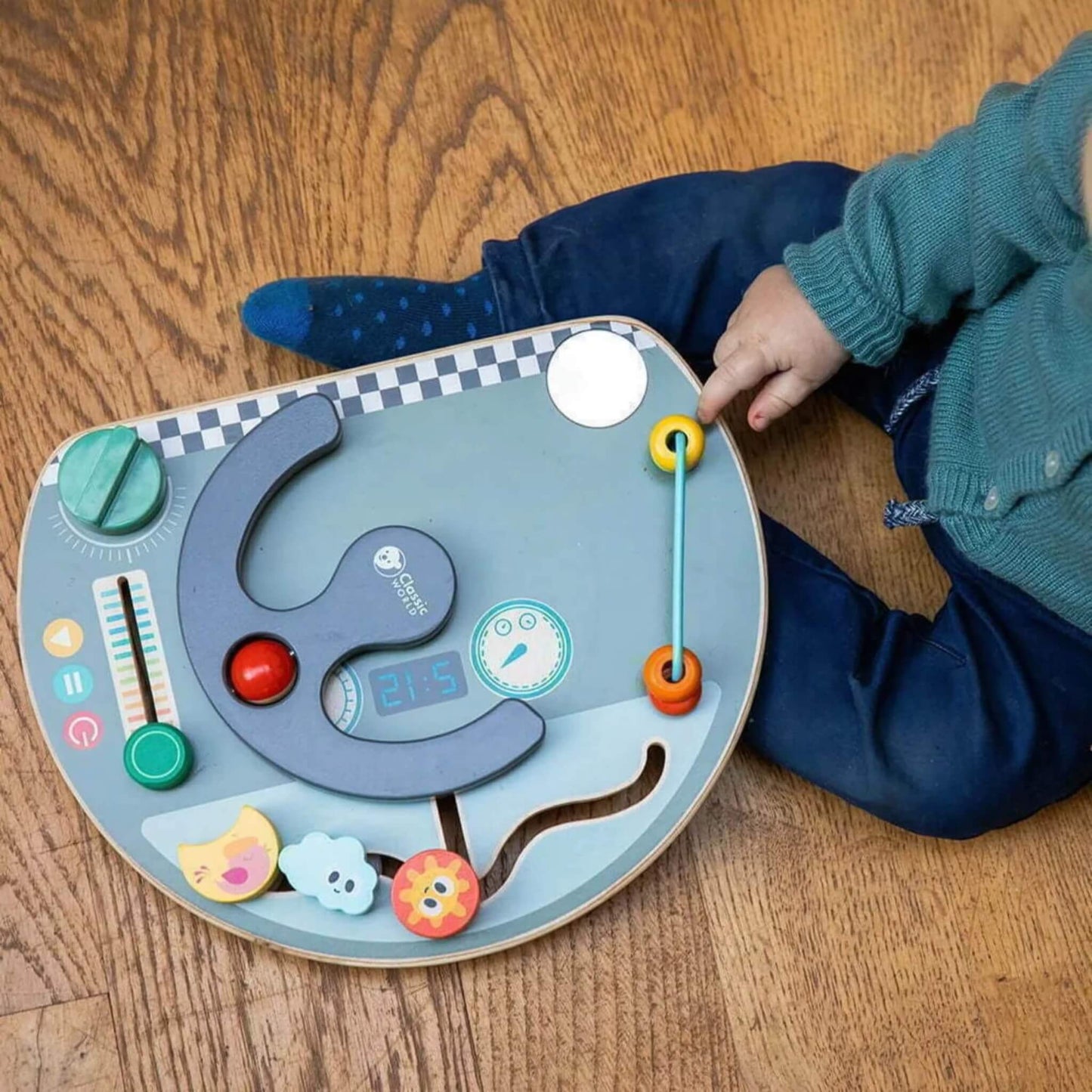 Overhead view of a toddler playing with a busy board on wooden flooring, touching the sliding beads.