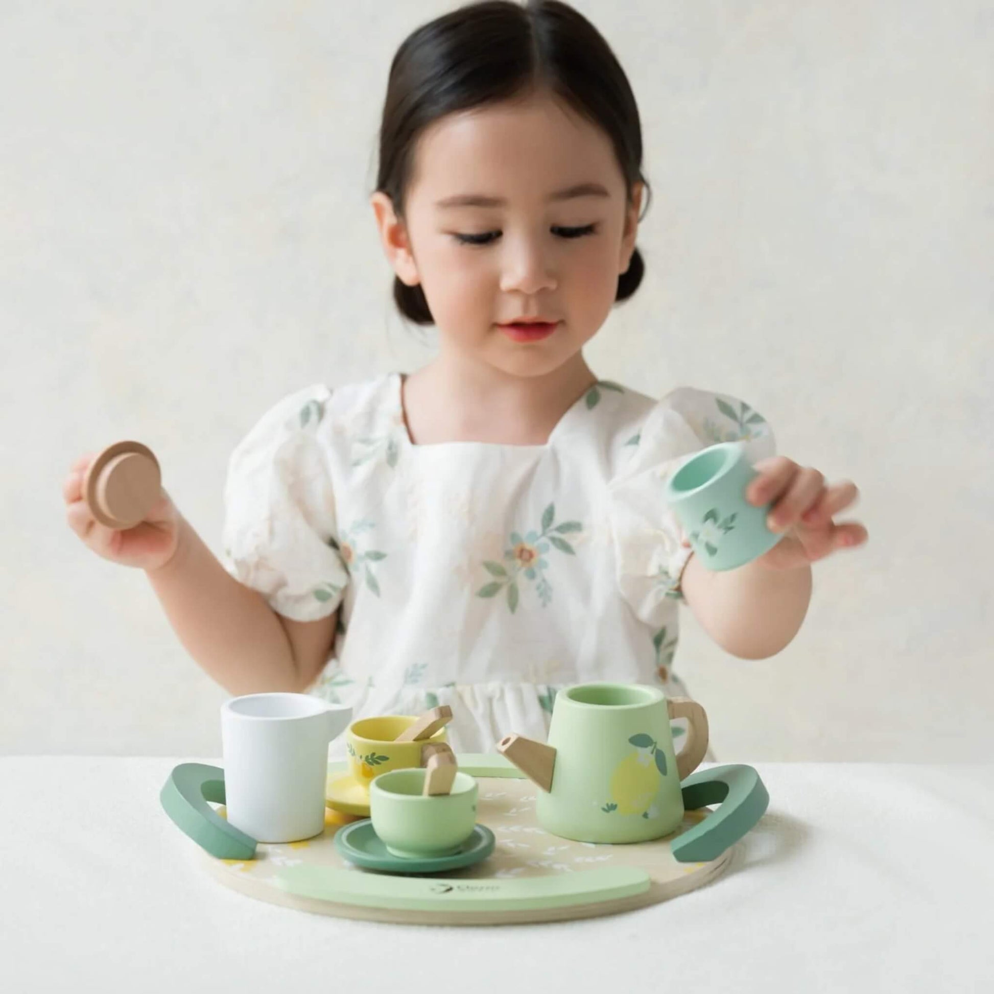 A young girl wearing a white floral dress plays with a wooden tea set on a table, pouring from a milk jug while holding a lid in her other hand.
