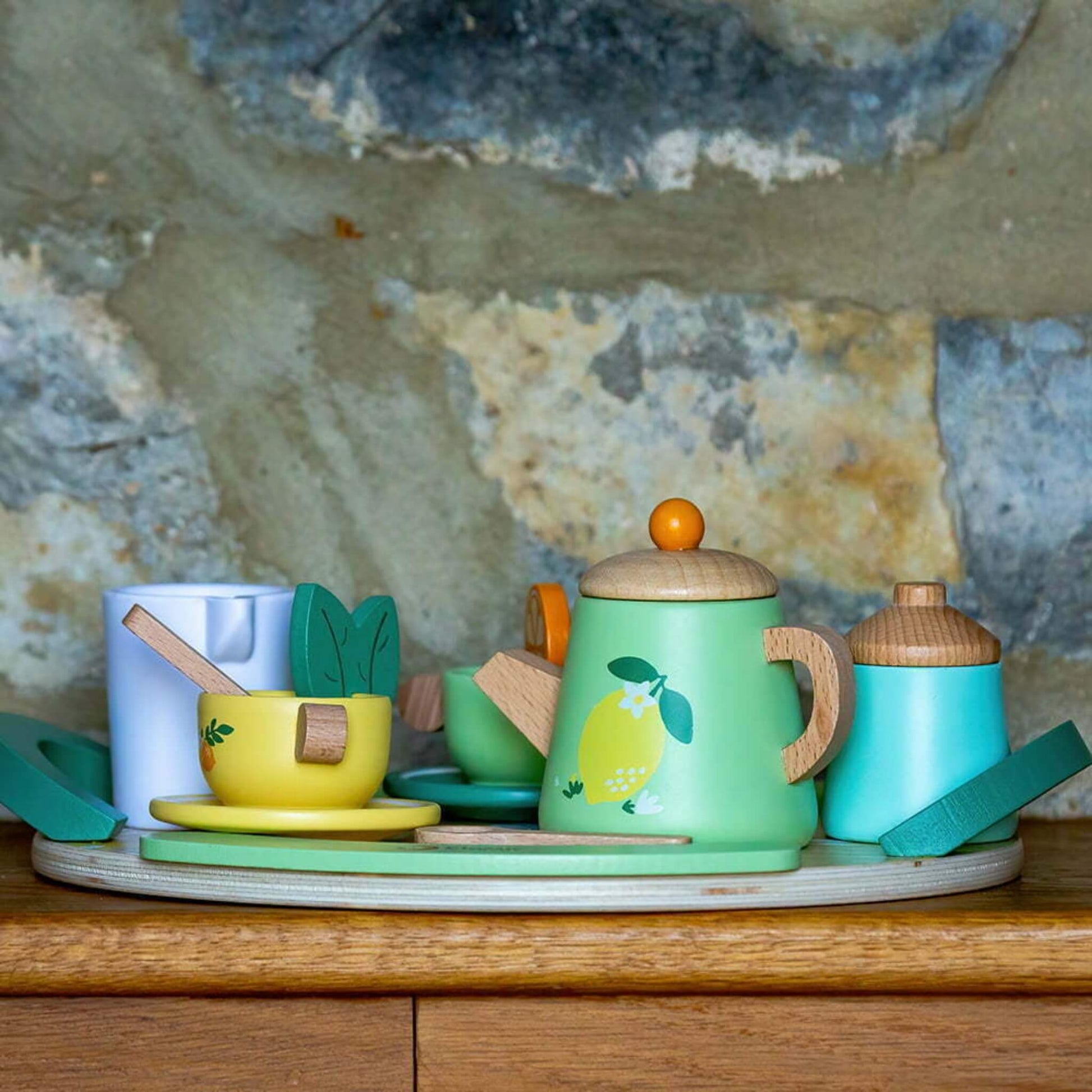 A wooden tea set with pastel colours displayed on a wooden surface in front of a rustic stone wall, showing a full arrangement of all play pieces.