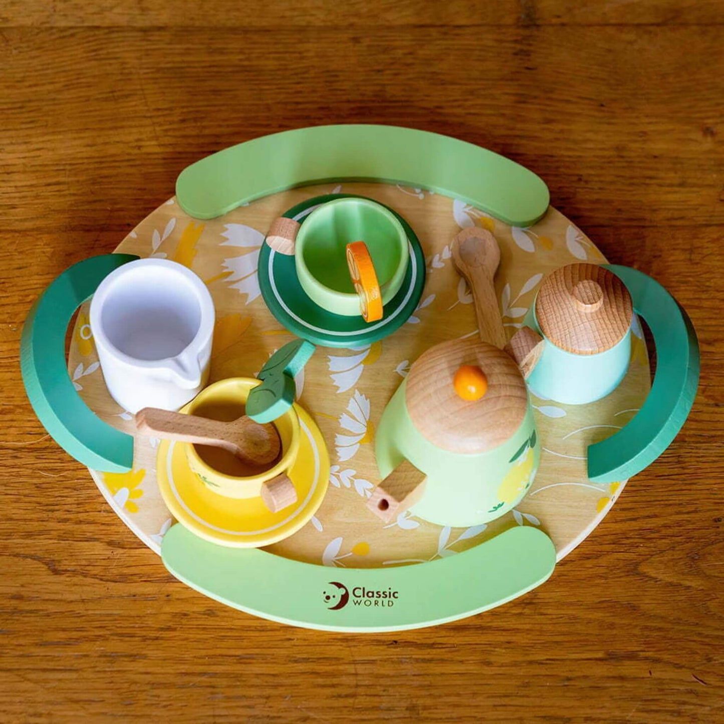 A top-down view of a wooden toy tea set arranged neatly on a floral tray, showing two teacups, teapot, jug, sugar bowl, spoons, and slice of lemon for pretend play.